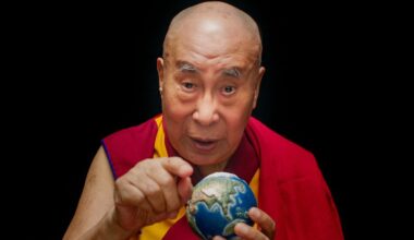 De Dalai Lama kijkt op deze scènefoto uit de film 'Wisdom of Happiness' (2024) recht in de camera. In zijn linkerhand heeft hij een wereldbol ter grootte van een tennisbal en met zijn rechterhand wijst hij naar de camera. Hij heeft een rood met geel kledingstuk aan.