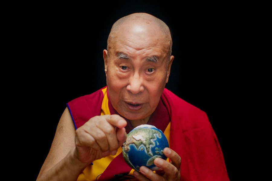 De Dalai Lama kijkt op deze scènefoto uit de film 'Wisdom of Happiness' (2024) recht in de camera. In zijn linkerhand heeft hij een wereldbol ter grootte van een tennisbal en met zijn rechterhand wijst hij naar de camera. Hij heeft een rood met geel kledingstuk aan.
