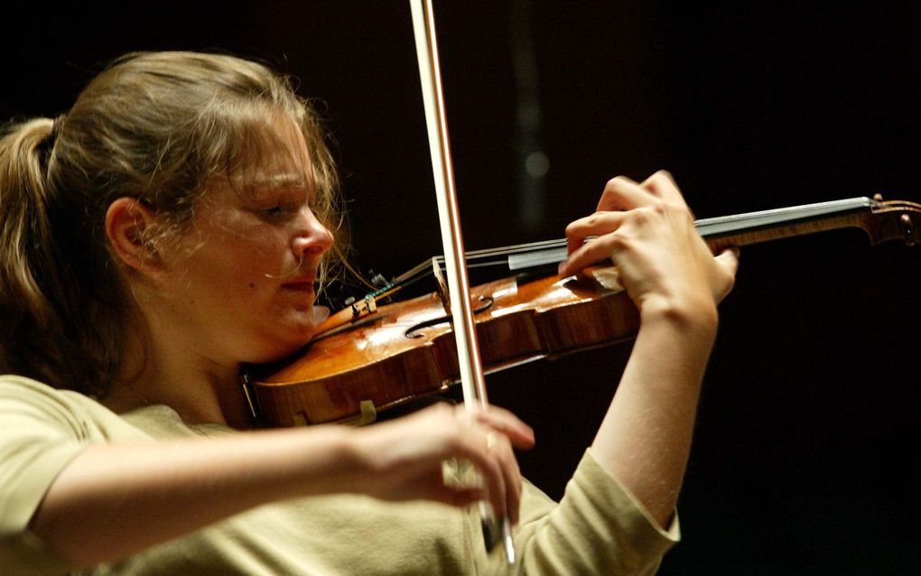 Een vrouw strijkt met een strijkstok over de snaren van de viool die ze viool onder haar kin heeft. Met haar linkerhand houdt ze de viool vast.