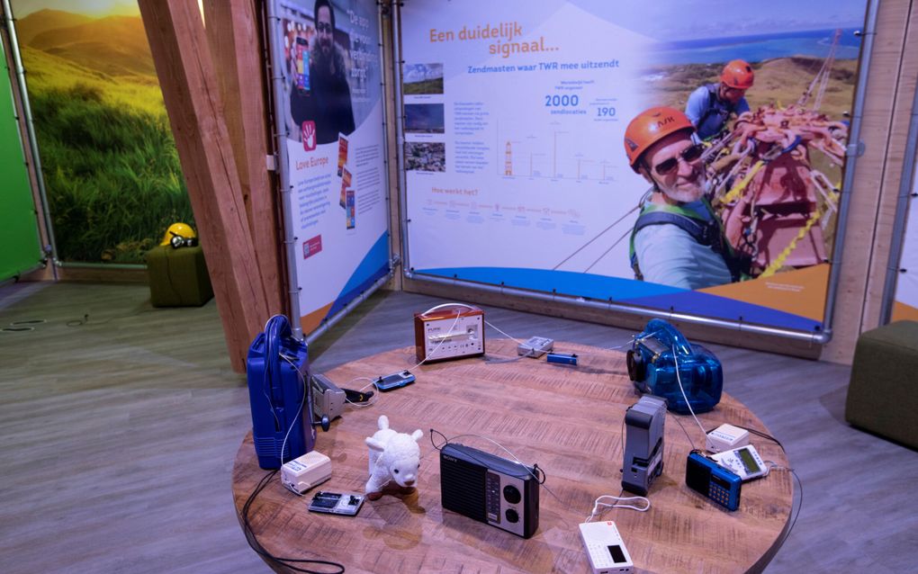 Tentoonstellingsruimte met een banner waarop mannen staan die werken aan een zendmast. Daarvoor staat een tafel waarop allerlei geluidsdragers liggen, zoals radio’s en andere apparatuur.
