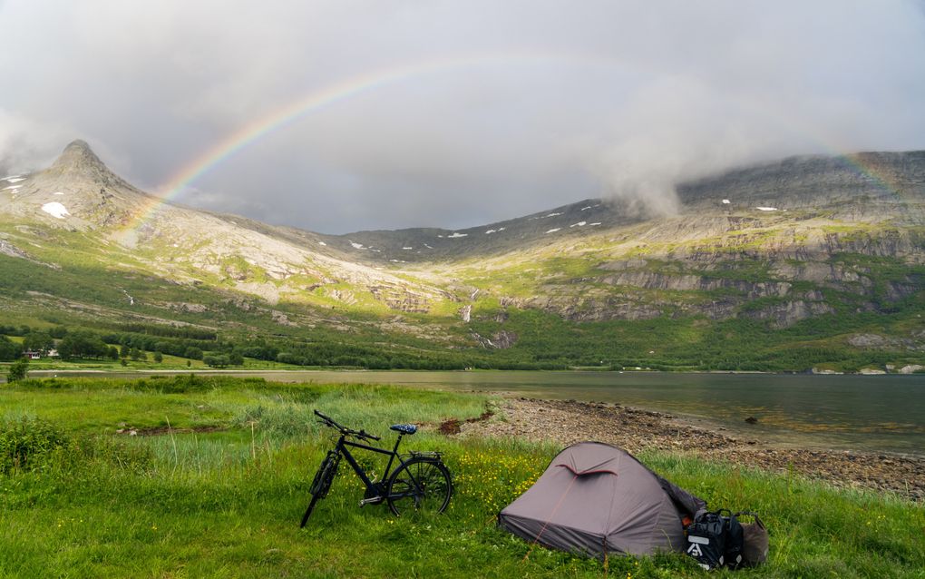 Een bruin tentje staat in groen gras aan het water. Tassen en fiets ernaast. De lucht is grijs, zon straalt over de heuvels, de regenboog is zichtbaar.
