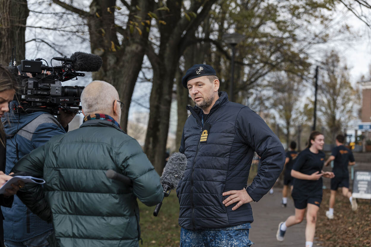 Kapitein-luitenant-ter zee (KLTZ) Erik Noordam staat de pers te woord terwijl op de achtergrond rekruten rennen.