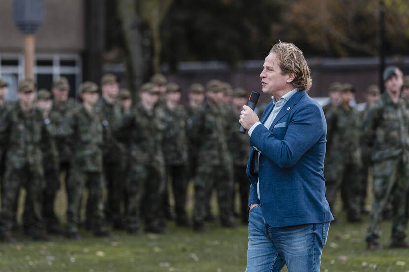 Staatssecretaris Gijs Tuiinman met microfoon in zijn hand. Op de achtergrond zijn vaag mensen in uniform te zien.