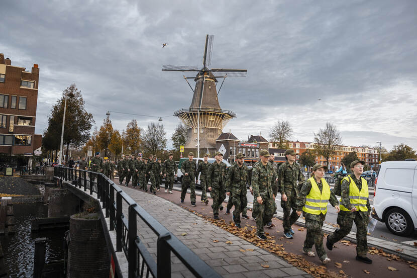 Jongeren in uniform lopen over een brug. Voorop 2 vrouwen met gele veiligheidshesjes aan. Op de achtergrond een molen. Ervoor vliegt een vogel.
