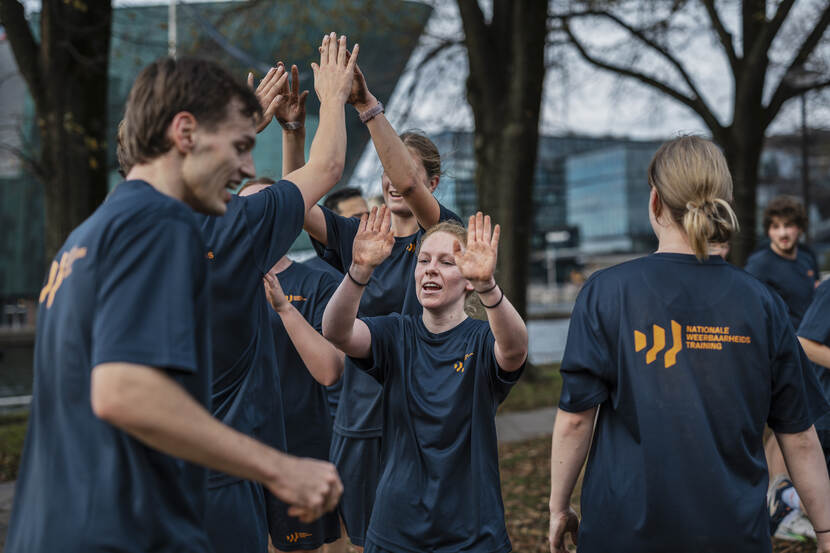 Een 8-tal jongeren met een T-shirt aan met de tekst Nationale Weerbaarheids Training erop. Met de armen omhoog klappen sommigen tegen de handen van een ander.