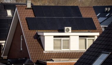 Een serie zonnepanelen ligt op een schuin dak, vlak boven de dakkapel van een huis dat in een woonwijk staat.