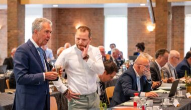 Tientallen mannen achter lange vergadertafels. De meeste zitten en kijken op hun laptop. Twee mannen staan en zijn in gesprek met elkaar.