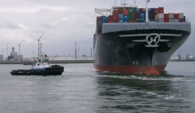 Containerschip in de haven, met heel veel gekleurde containers op het dek. Daarnaast een sleepboot.