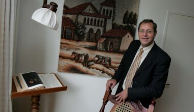 Man in donker pak met stropdas staat voor een wandkleed, leunend op een stoel.