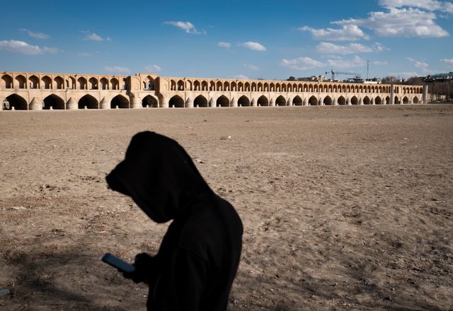 Een Iraanse man in Isfahan kijkt op 22 februari op zijn telefoon terwijl hij in de bedding van de opgedroogde Zayandehrivier staat. Naast hem doemt de bijna driehonderd meter lange Si-o-se Polbrug op, een stenen boogbrug uit de zestiende eeuw, die al eeuwenlang een toeristentrekker is voor de stad. De brug raakt beschadigd als de droogte aanhoudt.