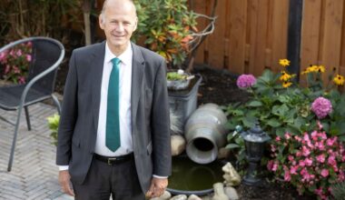 Man in pak, met stropdas, lachend. Hij staat in een tuin met erachter bloemen.