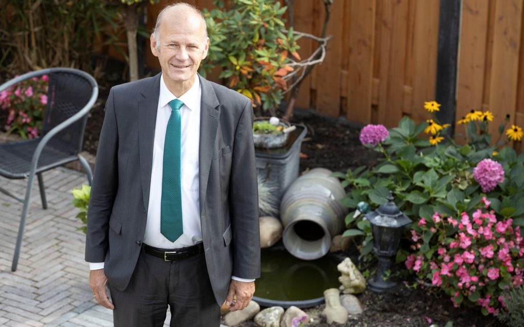 Man in pak, met stropdas, lachend. Hij staat in een tuin met erachter bloemen.