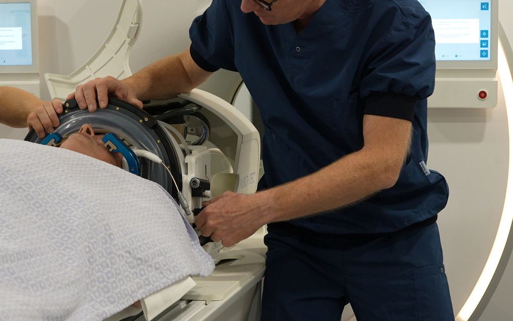 Een arts in blauwe kleding helpt een man die op een MRI-scanner ligt met een helm op zijn hoofd.