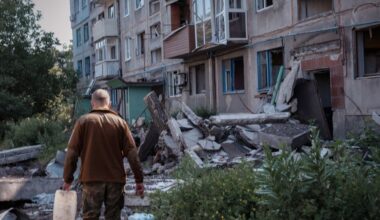 Een man met een bruine jas loopt met een jerrycan door een kapotgeschoten straat met kapotgeschoten flat