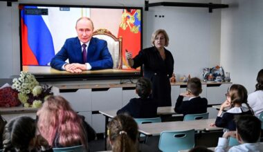 Kinderen kijken naar een digibord met daarop een man in pak op het scherm. 