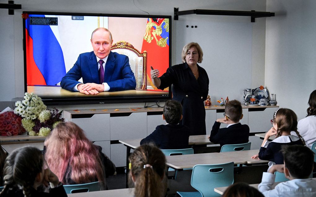 Kinderen kijken naar een digibord met daarop een man in pak op het scherm. 