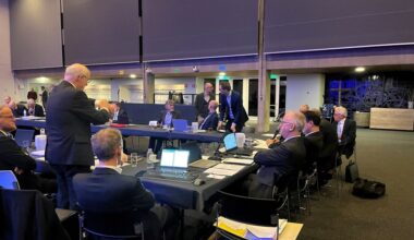 In een U-vorm geplaatste vergadertafels, met daarachter mannen in donkere pakken.