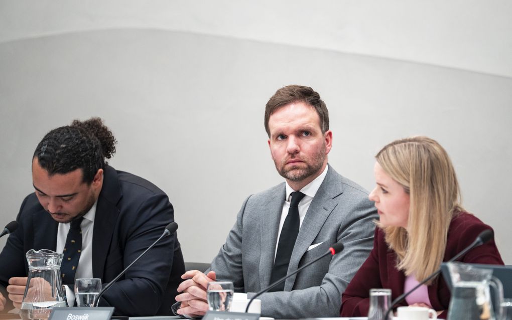 Twee mannen in pak en een vrouw zitten naast elkaar achter een tafel met daarop microfoons en naambordjes, in een commissiezaal van de Tweede Kamer.