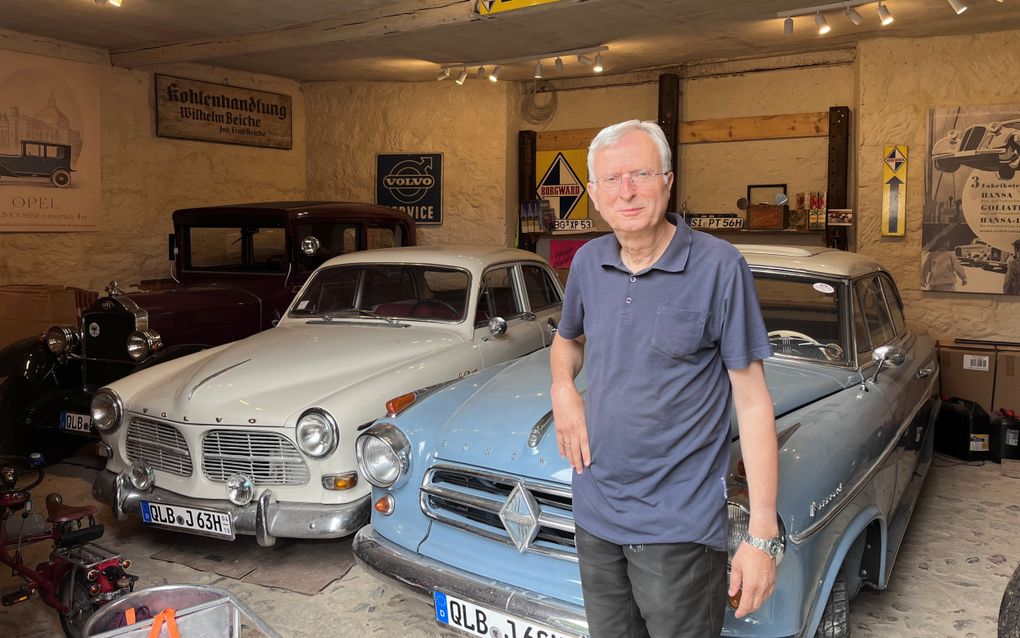 Een man staat voor drie klassieke auto’s. Op de voorgrond staan twee oude Volvo’s — een witte sedan en een lichtblauwe stationwagen — en achterin staat een vintageauto. Aan de muren hangen historische reclameborden van onder meer Volvo, Borgward en Opel.