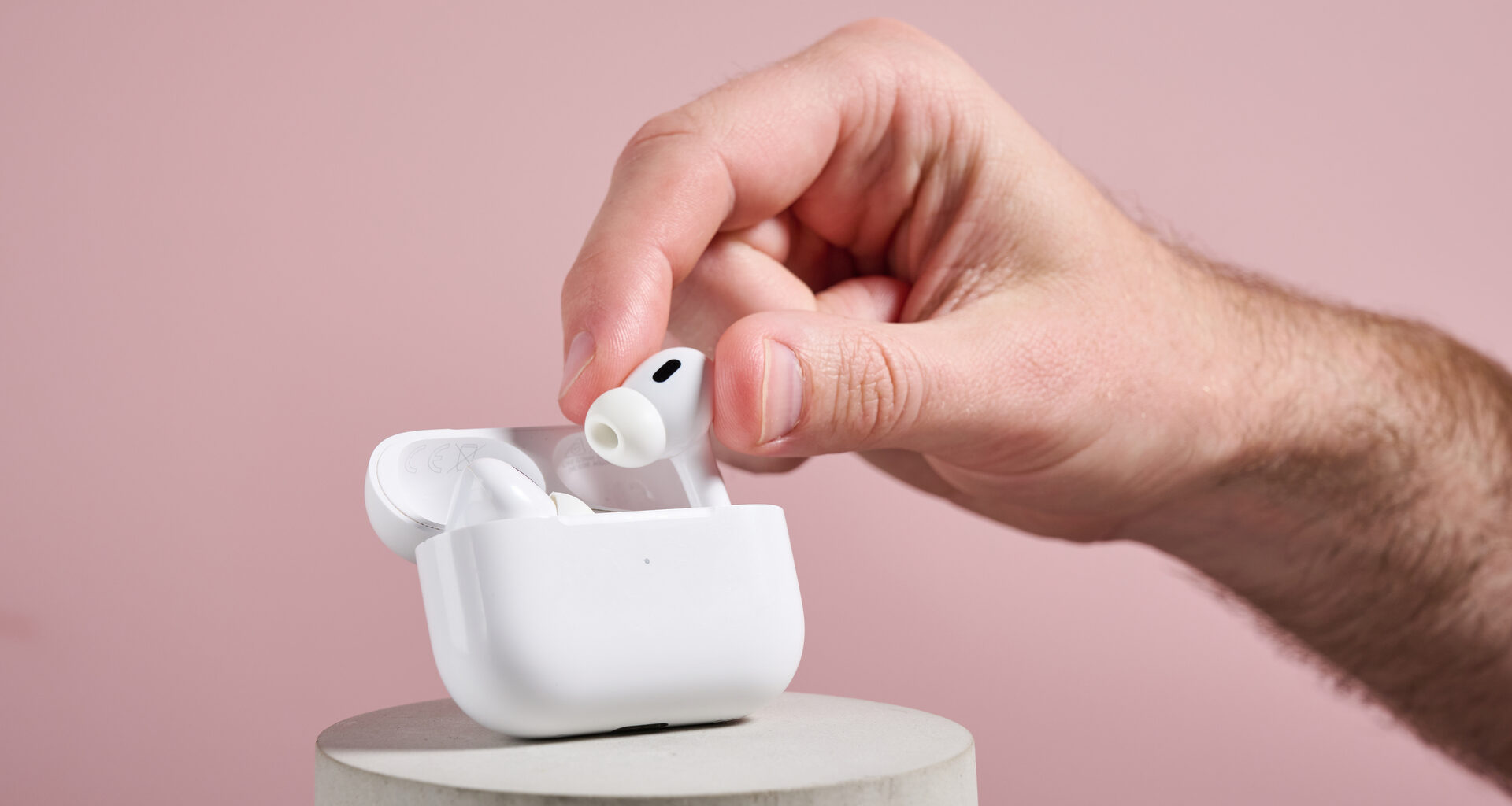 AirPods Pro 2 er tatt ut av ladeetuiet, som står på en grå søyle foran en rosa vegg.