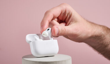 AirPods Pro 2 er tatt ut av ladeetuiet, som står på en grå søyle foran en rosa vegg.