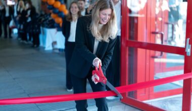 A smiling woman in a light sweater stands in an indoor hallway, a screen displaying "Peer Gynt-salen" behind her.
