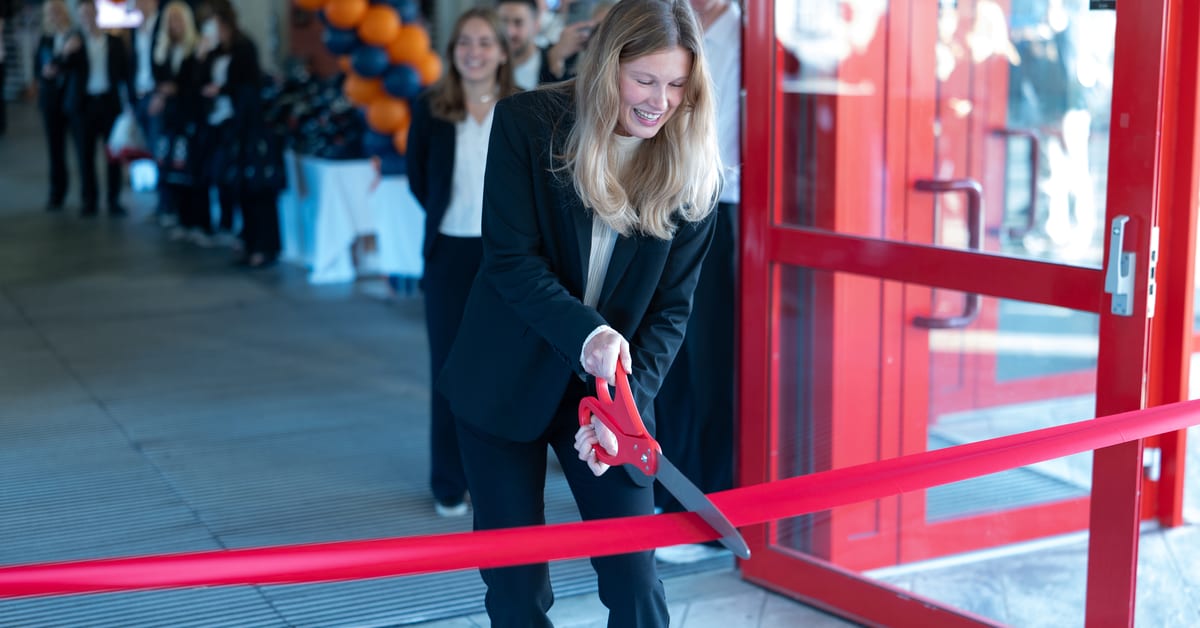 A smiling woman in a light sweater stands in an indoor hallway, a screen displaying "Peer Gynt-salen" behind her.
