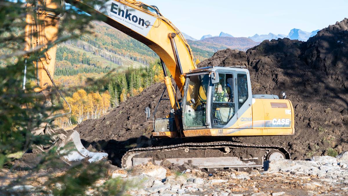 En gravemaskin/ bulldoser lemper matjorda i et naturområde med skog og fjell i bakgrunn