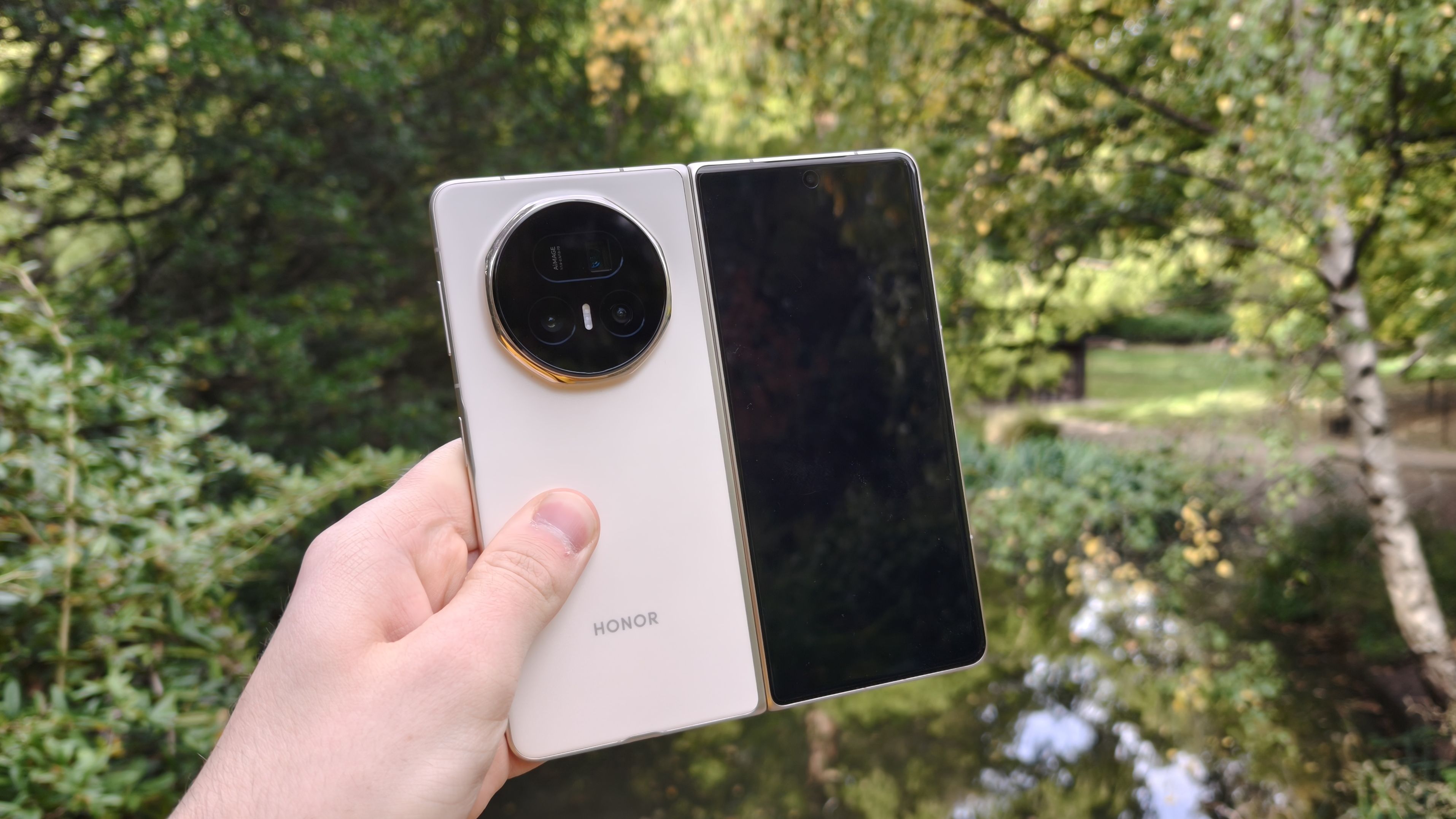 Honor Magic V5 i hvitt holdes foran en dam i en park.