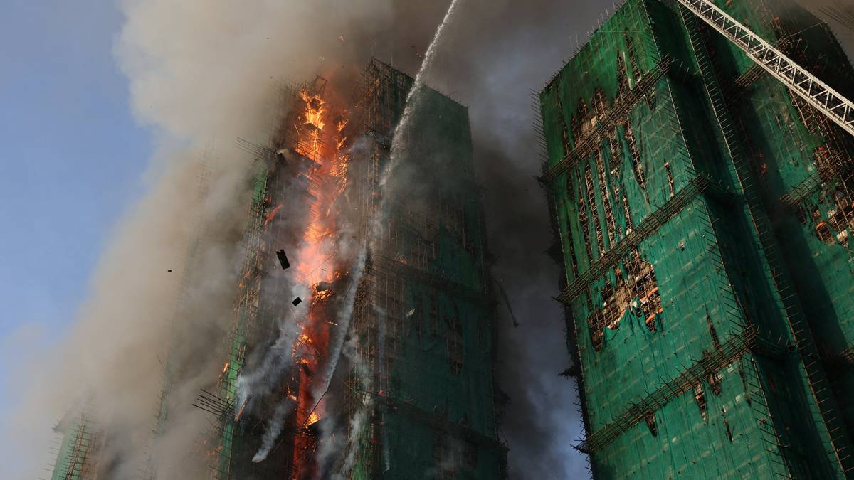 En høy bygning brenner kraftig, med flammer og tykk røyksky. Brannmenn sprøyter vann fra en høy stige for å slukke brannen. Bygningen har stillas og er delvis dekket med grønt nettingmateriale. Omgivelsene er uklare på grunn av røyken.
