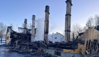 En bygning står utbrent med kun rester av vegger og pipestokker synlige. Røyk stiger fra ruinen, og det er snø på bakken rundt. Bakgrunnen viser en intakt hvit bygning og trær, som gir kontrast til det ødelagte området. Politibånd er plassert rundt stedet, som indikerer at området er avsperret.