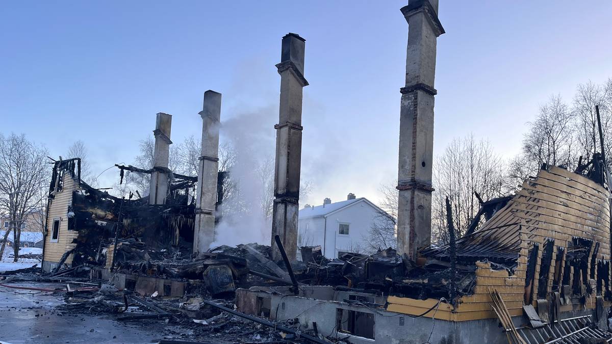 En bygning står utbrent med kun rester av vegger og pipestokker synlige. Røyk stiger fra ruinen, og det er snø på bakken rundt. Bakgrunnen viser en intakt hvit bygning og trær, som gir kontrast til det ødelagte området. Politibånd er plassert rundt stedet, som indikerer at området er avsperret.