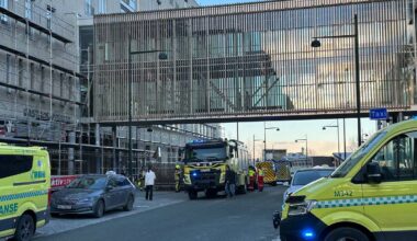 Utrykningskjøretøy fra brannvesenet og ambulanse er samlet ved en byggeplass. Flere personer i arbeidstøy står i nærheten. En sykebil er parkert på fortauet, og det er en tydelig byggeaktivitet i området. Omgivelsene inkluderer en, så halvbygget bygning med stillas og en heis.