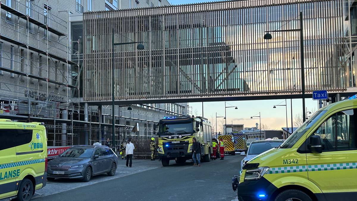 Utrykningskjøretøy fra brannvesenet og ambulanse er samlet ved en byggeplass. Flere personer i arbeidstøy står i nærheten. En sykebil er parkert på fortauet, og det er en tydelig byggeaktivitet i området. Omgivelsene inkluderer en, så halvbygget bygning med stillas og en heis.