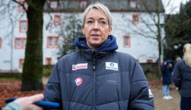 En person i en blå boblejakke står foran en bygning med røde vinduer. Det er høst og bakken er dekket av blader. En annen person er synlig i bakgrunnen og ser ut til å bruke en mobiltelefon. Omgivelsene ser ut til å være en utendørs arena eller et offentlig område.