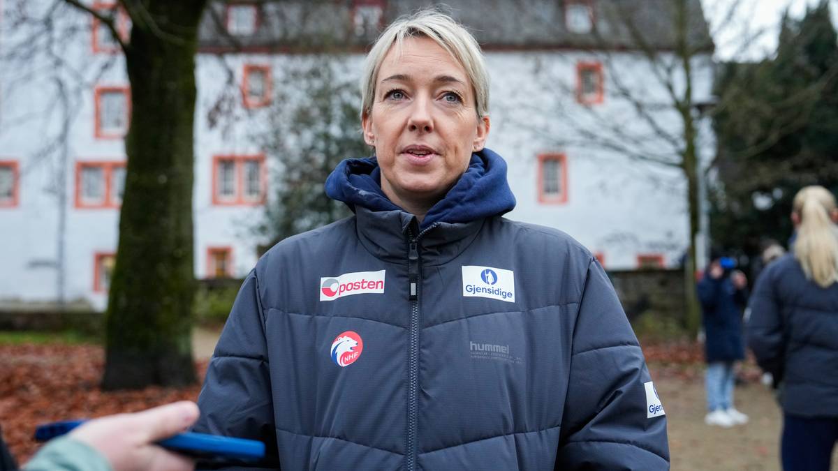 En person i en blå boblejakke står foran en bygning med røde vinduer. Det er høst og bakken er dekket av blader. En annen person er synlig i bakgrunnen og ser ut til å bruke en mobiltelefon. Omgivelsene ser ut til å være en utendørs arena eller et offentlig område.