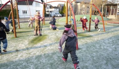 Flere barn leker på en lekeplass, hvor noen svinger og andre løper omkring. Bakgrunnen viser huse i nabolaget. Det er et trevlet underlag, og barnas klær varierer i farger og stil. Sollyset skaper lange skygger på bakken.