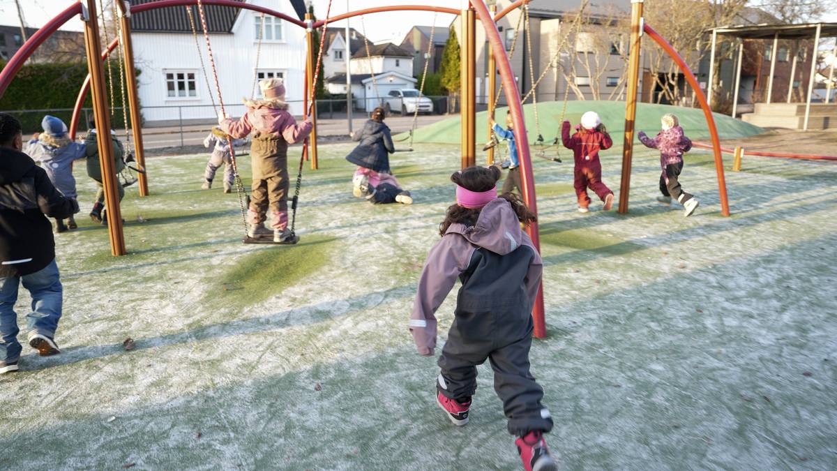 Flere barn leker på en lekeplass, hvor noen svinger og andre løper omkring. Bakgrunnen viser huse i nabolaget. Det er et trevlet underlag, og barnas klær varierer i farger og stil. Sollyset skaper lange skygger på bakken.