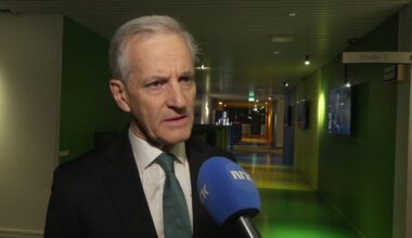 En mann i dress holder en mikrofon med NRK-logo. Omgivelsene er et fargerikt, opplyst gangområde. I bakgrunnen er det flere TV-skjermer og en dør merket "Studio 3". Belysningen er dempet og moderne.