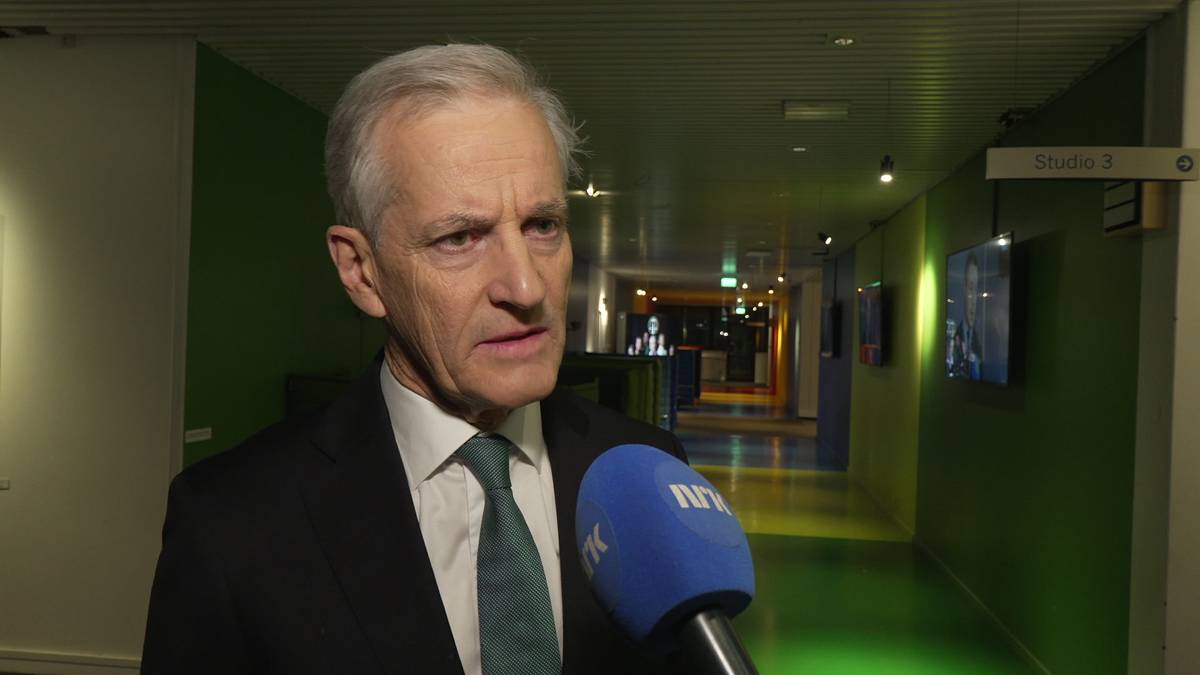 En mann i dress holder en mikrofon med NRK-logo. Omgivelsene er et fargerikt, opplyst gangområde. I bakgrunnen er det flere TV-skjermer og en dør merket "Studio 3". Belysningen er dempet og moderne.