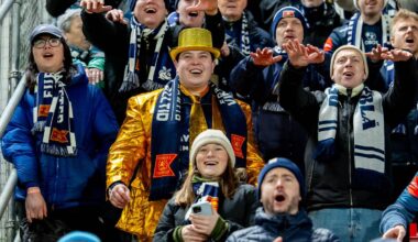 En gruppe entusiaster heier på et idrettsarrangement. Noen bærer skjerf med klubblogoer, mens en person i en gylden jakke skiller seg ut. Publikum viser engasjement med hendene hevet. Omgivelsene inkluderer tribuner og en metallgjerde.