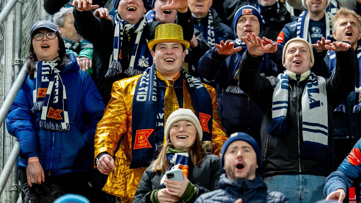 En gruppe entusiaster heier på et idrettsarrangement. Noen bærer skjerf med klubblogoer, mens en person i en gylden jakke skiller seg ut. Publikum viser engasjement med hendene hevet. Omgivelsene inkluderer tribuner og en metallgjerde.