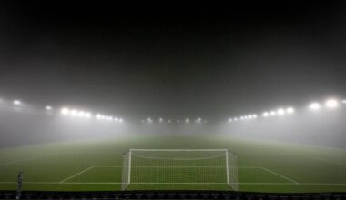 Et fotballstadion er delt inn i flere seksjoner, men er nesten helt dekket av tåke. Kun målet og noen lys er synlige, og banen ser ut til å være av kunstgress. Lyskasterne står lysende over den tomme arenaen. Ingen personer er synlige i bildet.