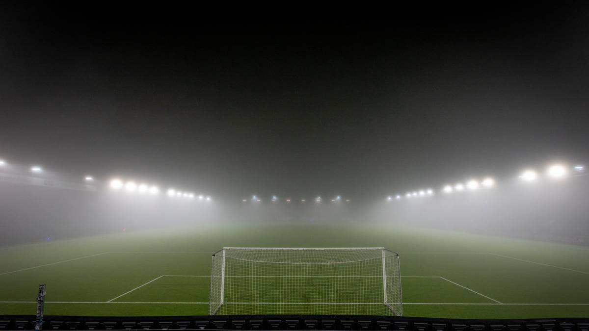 Et fotballstadion er delt inn i flere seksjoner, men er nesten helt dekket av tåke. Kun målet og noen lys er synlige, og banen ser ut til å være av kunstgress. Lyskasterne står lysende over den tomme arenaen. Ingen personer er synlige i bildet.