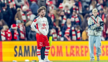 To fotballspillere står på banen under en kamp, mens publikum følger med i bakgrunnen. Den ene spilleren har på seg rød og hvit drakt, mens den andre er iført en jakke. Det er mange tilskuere på tribunen kledd i lagets farger. Det er lys fra stadion og reklamebannere synlige i området.