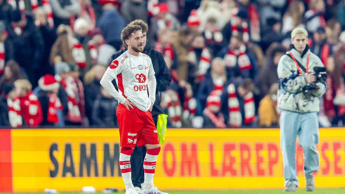 To fotballspillere står på banen under en kamp, mens publikum følger med i bakgrunnen. Den ene spilleren har på seg rød og hvit drakt, mens den andre er iført en jakke. Det er mange tilskuere på tribunen kledd i lagets farger. Det er lys fra stadion og reklamebannere synlige i området.