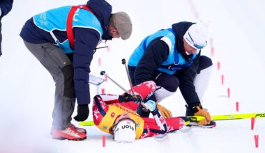 To personer i blå vest hjelper en skiskytter som ligger på bakken. Utøveren har på seg rød drakt med sponsorlogoer. Omgivelsene er snødekte, med spor etter ski synlig i bakgrunnen. Det er offisielle markeringer i snøen rundt utøveren.