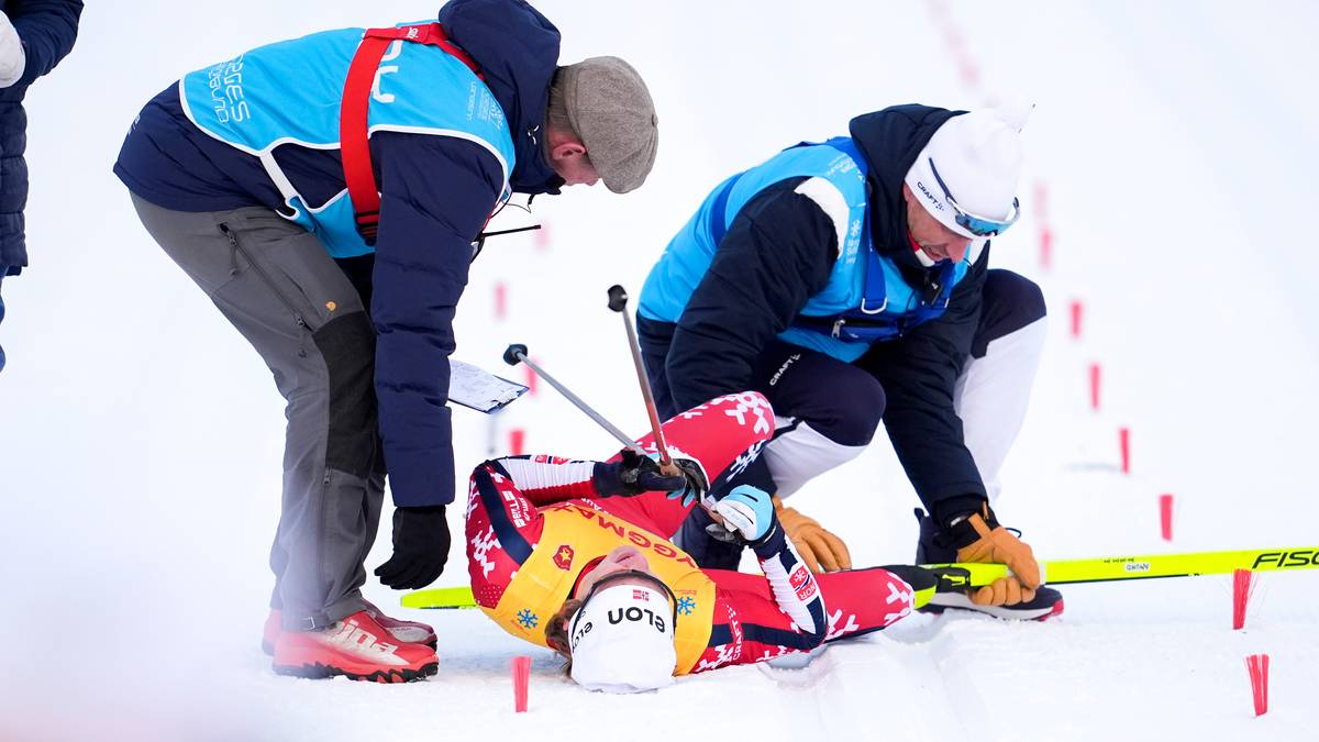 To personer i blå vest hjelper en skiskytter som ligger på bakken. Utøveren har på seg rød drakt med sponsorlogoer. Omgivelsene er snødekte, med spor etter ski synlig i bakgrunnen. Det er offisielle markeringer i snøen rundt utøveren.