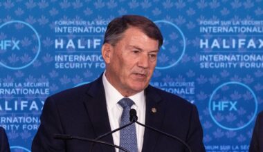 Tre menn står ved en talerstol under Halifax International Security Forum. Den midterste mannen taler, mens de to andre står til venstre og høyre. Det er en felles bakgrunn med teksten "Halifax International Security Forum". Mennene er kledd i formelle klær og ser ut til å være engasjert i diskusjonen.