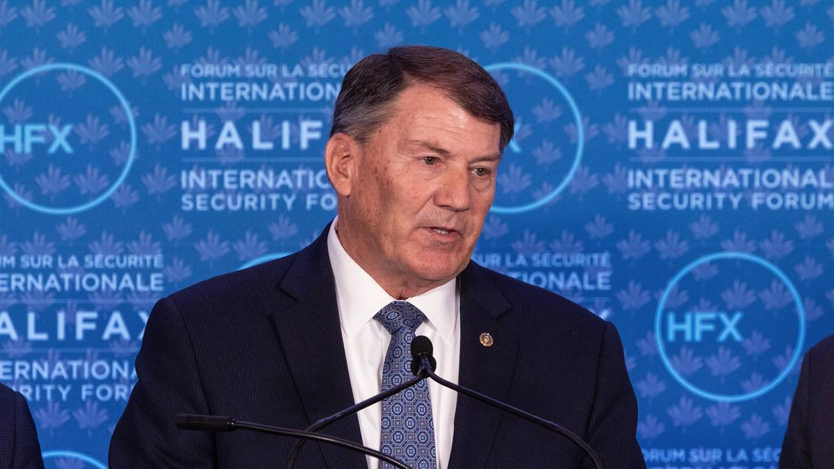 Tre menn står ved en talerstol under Halifax International Security Forum. Den midterste mannen taler, mens de to andre står til venstre og høyre. Det er en felles bakgrunn med teksten "Halifax International Security Forum". Mennene er kledd i formelle klær og ser ut til å være engasjert i diskusjonen.